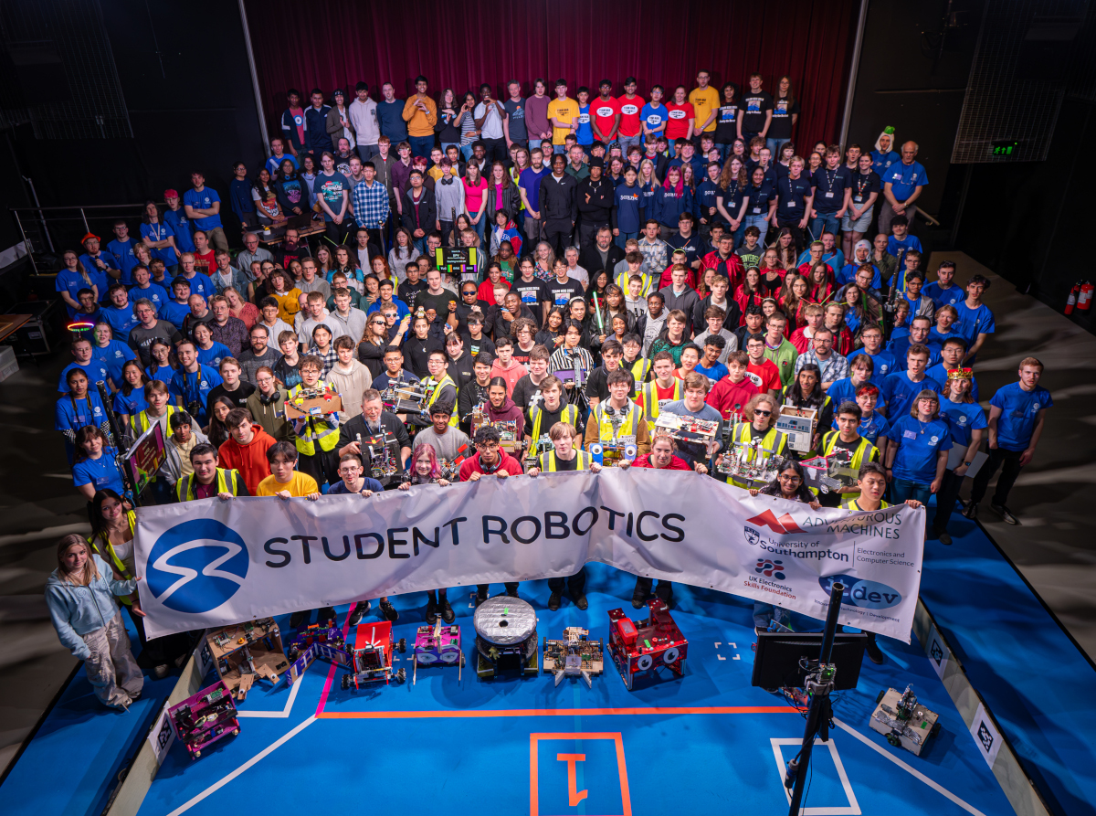 
            The competitors and volunteers from the SR2024 competition, standing in and around the arena.
            At the front the competitors hold a Student Robotics banner, with a selection of robots on the ground in front of them.
            The volunteers are all wearing the customary blue tee-shirts identifying them as such.
          